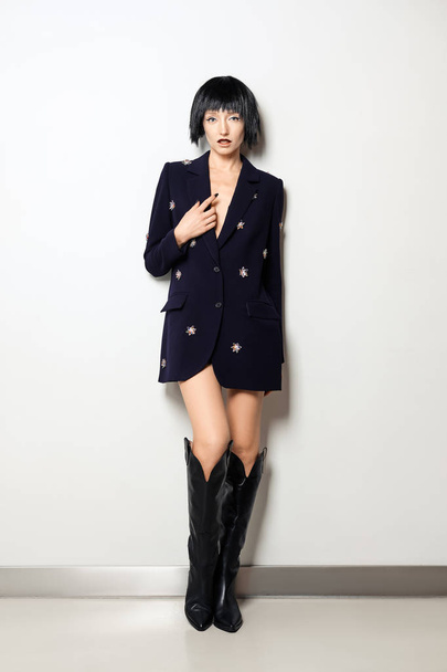 Fashion model with black bob haircut posing near the wall in blue jacket with gems decor and in wide high boots - Φωτογραφία, εικόνα