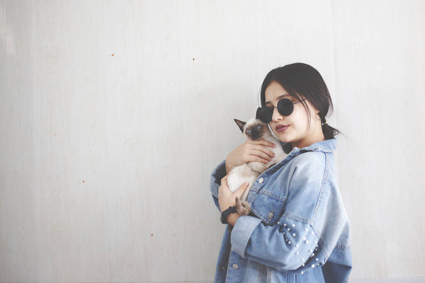 Молодая привлекательная женщина обнимает киску в руках. Милая и гламурная девушка в модных солнцезащитных очках позирует со своей сиамской кошкой
 - Фото, изображение