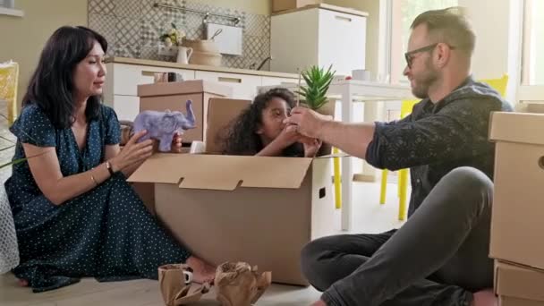 Boldog vegyes etnikum mosolygó család kisfiúval és kislánnyal dobozok kipakolása együtt ül a kanapén a modern hangulatos nappaliban, boldog szülők játszanak a gyerekekkel kipakolás - Felvétel, videó