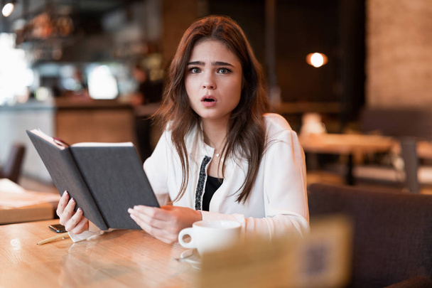 young beautiful woman in white jacket checking her planner working outside office drinking hot coffee in the trendy cafe looking confused multitasking modern businesswoman - Fotografie, Obrázek