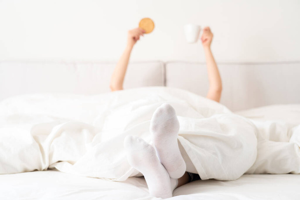 Female hand holding cup of coffee and cracker from under a blanket in bed. Woman waking up in the morning - Fotó, kép