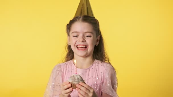 Happy beautiful girl in party cap blowing out candle on yellow background. - Séquence, vidéo
