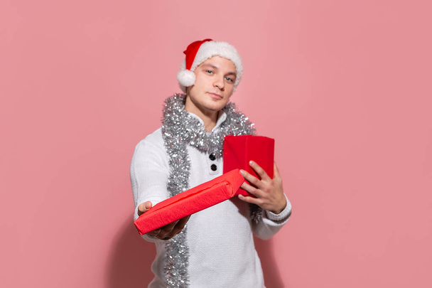Hombre guapo en un suéter blanco y un sombrero de Santa Claus rojo sosteniendo regalos de Navidad en cajas rojas aisladas sobre fondo rosa. Feliz Navidad y Feliz Año Nuevo
 - Foto, imagen