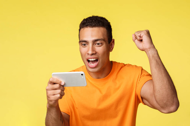 Joyful excited young handsome man in orange t-shirt, raise hand up fist pump as rooting for favorite team, holding smartphone horizontally, watching football on mobile device, yellow background - Fotografie, Obrázek