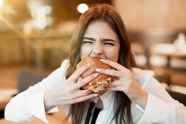 young woman in white stylish blouse biting with appetite fresh tasty burger during lunch in trendy cafe feeling ultimate hunger eating outside - Foto, Imagem