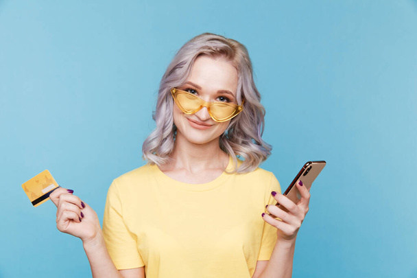 Жінка в окулярах з кредитною карткою і мобільний ізольовані над синьою студією
. - Фото, зображення