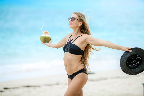 Woman in a black swimsuit on the beach drinks coconut juice from fresh coconut - Zdjęcie, obraz