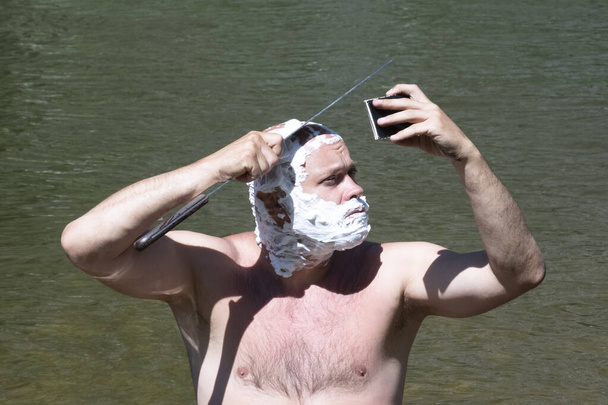 Kopf und Gesicht eines Mannes sind mit Seifenschaum bedeckt. Ein brutaler Typ sieht sich selbst im Spiegel an und rasiert sich seine Glatze mit einem scharfen Machetenmesser auf Wassergrund. Lebensstil der Reisenden. - Foto, Bild