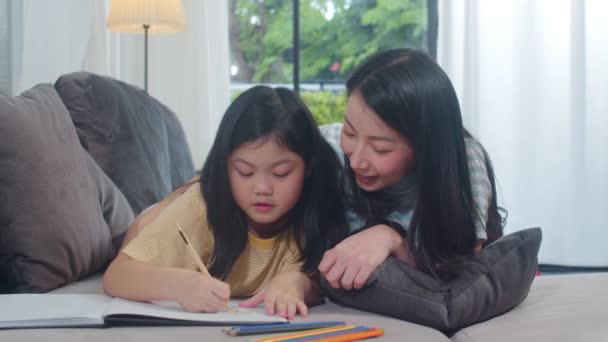 Asian middle-aged woman teach her daughter do homework and drawing at home. Lifestyle mother and kid happy fun spend time together in living room in modern home in the evening concept. - Footage, Video