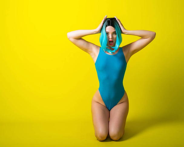 Beautiful girl in a blue wig and a bright bikini posing on a yellow background. Woman with artificial hair and a swimsuit emphasizing the buttocks. - Photo, image
