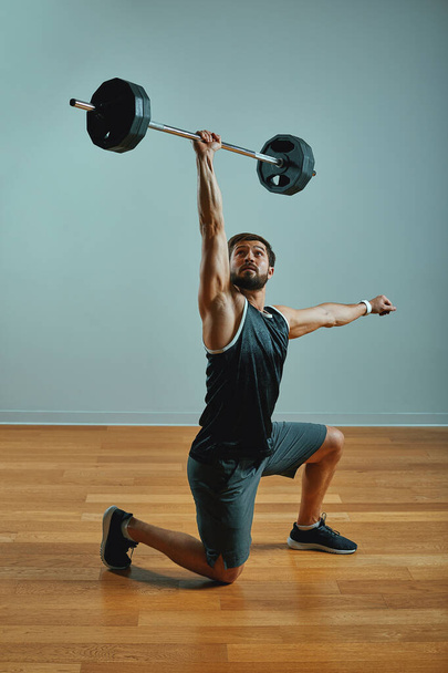Retrato de un joven musculoso súper en forma haciendo ejercicio en el gimnasio con barra de pesas sobre fondo gris, copyspace
. - Foto, imagen