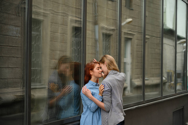 Same-sex relationships. Portrait of two young lesbians passionately hugging and standing against a mirror wall. Loving couple of gay women standing on the street and showing feelings. - Фото, изображение