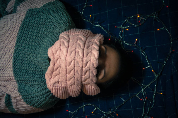 The girl in the winter gray-pink sweater lies on a plaid with a garland covering her face with pink scarf - Fotoğraf, Görsel