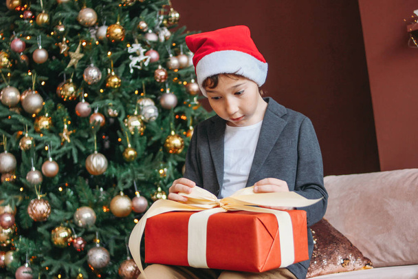 Atractivo chaval divertido con pelo oscuro en caja de regalo abierta del sombrero de santa sobre fondo del árbol de Navidad
 - Foto, Imagen