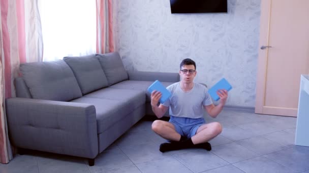 Weak nerd man is doing exercises for hand biceps with yoga blocks instead of dumbbells sitting on the floor at home. Sport humor concept. - 映像、動画