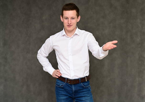 Concept of a pleasant young man manager talking to the camera. Photo Portrait of a secretary guy on a gray background in blue jeans and a white shirt in various poses. - Foto, Bild