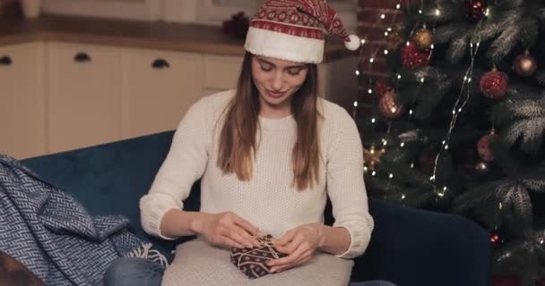 Close Up of Attractive Girl Wearing Sweater and Christmas Hat Sitting on the Sofa near Christmas Tree Finalize Small Christmas Gift Bandaging Tape and Tied in a Bow at Home Background - Filmati, video