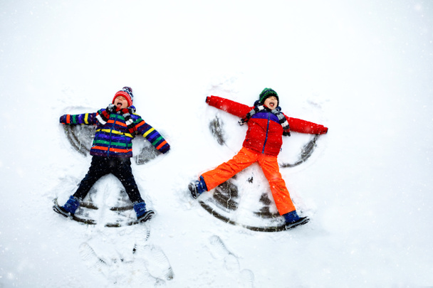 Dos hermanos pequeños niños con ropa de invierno de colores haciendo ángel de nieve, acostado en la nieve. Activo ocio al aire libre con niños en invierno. Hermanos felices con sombrero caliente, guantes, moda de invierno. - Foto, imagen