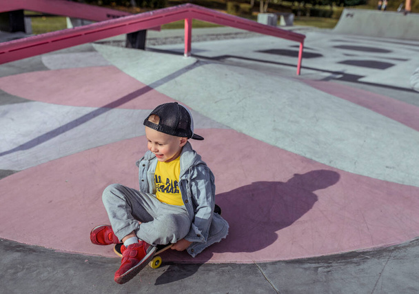 Little boy 4-5 years old, in autumn summer city on sports field, learning to ride a skateboard, happy smiling, free space for text. Casual wear denim cap, sneaker pants. - Photo, Image