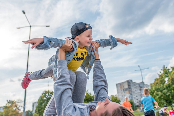 Mom woman with little boy son 4-5 years old, play airplane, have fun, laugh and enjoy rest, in summer city park site. Casual wear denim cap, sneaker pants. Background kids teens. - Photo, image