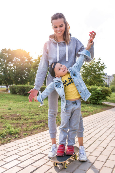 Young mother woman with little boy son of 4-5 years old, Support help balance training. Emotions of joy fun learns ride skateboard, summer autumn in city park playground. Skateboard, casual wear. - Foto, Bild