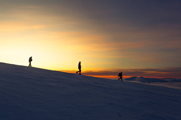 Három ember ereszkedik le a hegyről napnyugtakor télen, meredek lejtőn, a távolban. - Fotó, kép