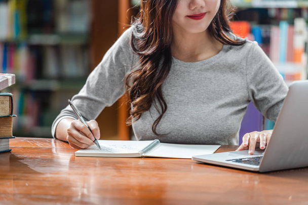 Closeup ázsiai fiatal diák kézzel írás házi feladatot, és a technológiát használó laptop a könyvtárban az egyetemi vagy kolléga különböző könyvet, és helyhez kötött a könyv alatt shelf háttér, vissza az iskolába - Fotó, kép