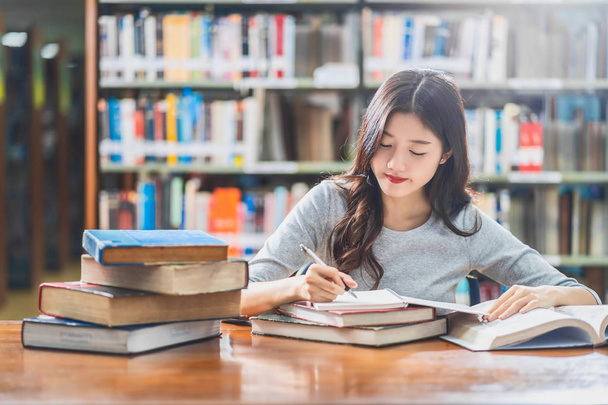 Mladý asijský student v příležitostném obleku čtení a práce s domácími úkoly v knihovně univerzit nebo kolektivu s různými knižní a stojící na dřevěném stole nad knižní tapetu, zpět do školy - Fotografie, Obrázek