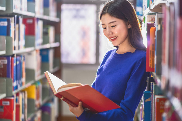 カジュアルなスーツを着たアジアの若い学生が立ち上がり、大学の図書館の本棚で本を読んだり、様々な本の背景を持つコラージュ、学校の概念に戻る - 写真・画像