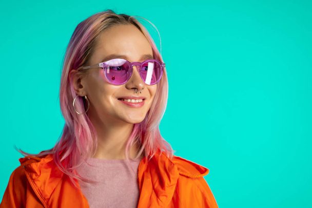 Cheerful odd girl in blue studio. Smiling woman with nose piercing, sunglasses. Vibrant colorful footage.  - Valokuva, kuva