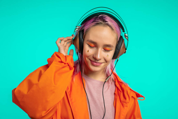 Pretty girl with pink hair enjoys listening to music. Hipster woman with extraordinary appearance in orange raincoat on blue background. - Photo, image