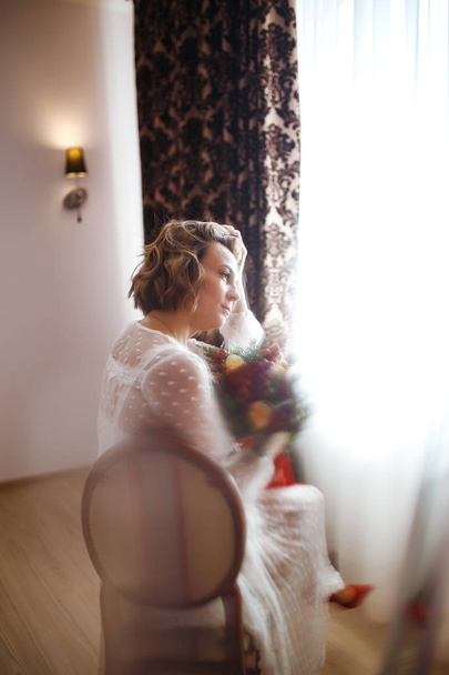 Красивая невеста со свадебным букетом цветов в белом роскошном платье готовится к свадьбе. Утренняя подготовка. Невеста со свадебным макияжем и прической. В день свадьбы. Брак
. - Фото, изображение