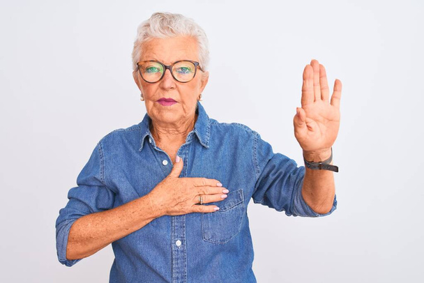 Старшая седовласая женщина в джинсовой рубашке и очках на изолированном белом фоне, ругаясь рукой на груди и открытой ладони, давая клятву верности
 - Фото, изображение