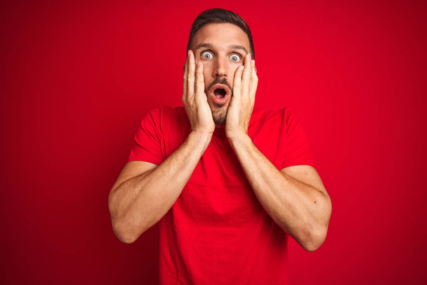 Mladý pohledný muž na sobě ležérní tričko přes červené izolované pozadí strach a šok, překvapení a užaslý výraz s rukama na tváři - Fotografie, Obrázek