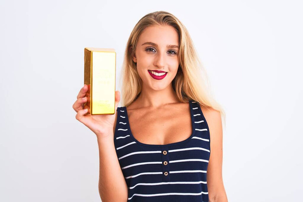 Νεαρή όμορφη ξανθιά γυναίκα που κρατά ράβδους χρυσού στέκεται πάνω από απομονωμένο λευκό φόντο με ένα χαρούμενο πρόσωπο στέκεται και χαμογελά με αυτοπεποίθηση χαμόγελο που δείχνει τα δόντια - Φωτογραφία, εικόνα
