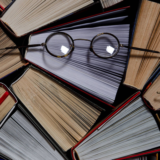Many multicolored thick open books stand on a dark background. On the books are old round glasses - Photo, Image
