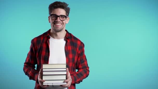 European student in red plaid shirt on blue background in studio holds stack of university books from library. Copy space. Guy smiles, he is happy to graduate. - 映像、動画