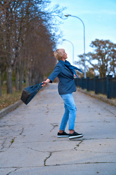 Счастливая блондинка ходит и крутится по осеннему городскому парку, одетая в синие джинсы и куртку
 - Фото, изображение