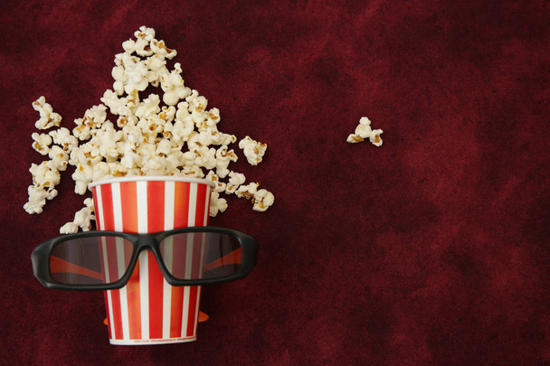 Popcorn im Pappbecher und Popcorn fliegen mit Filmgläsern auf rotem Hintergrund mit Platz für Text heraus. flache Lage, Draufsicht. Hintergrund, Muster, Karte. das Konzept des Kinos. - Foto, Bild