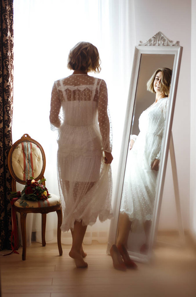 Bride in elegant white dress looking in the mirror. The bride smiles and poses in front of the mirror. Beautiful woman with professional make up and hair style. Morning of the bride preparations.  - Foto, Bild