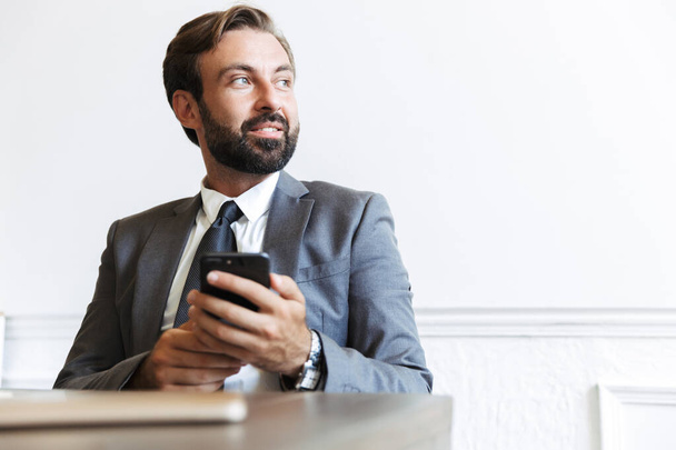 Image of bearded satisfied businessman typing on cellphone and looking aside while working in office - Photo, Image