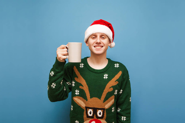 Portret van een lachende jongeman in een groene warme trui en hoed van de kerstman, staande tegen een blauwe achtergrond met een kopje warme drank in de hand, kijkend in de camera en glimlachend. - Foto, afbeelding