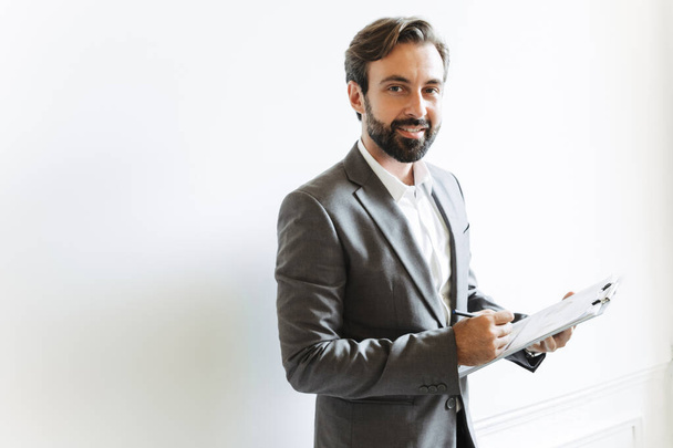 Image of happy successful businessman holding clipboard and writing down notes while working in office - Φωτογραφία, εικόνα