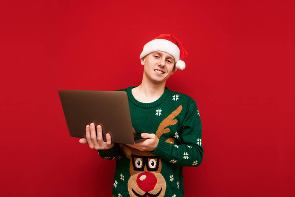 Счастливый молодой человек фрилансер с ноутбуком в руке изолированы на красном фоне, глядя в камеру и улыбаясь. Портер рождественского мальчика с ноутбуком на красном фоне. Копирование пространства
 - Фото, изображение