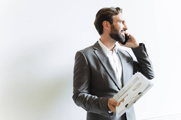 Image of handsome caucasian businessman talking on cellphone and holding clipboard while working in office - Fotografie, Obrázek