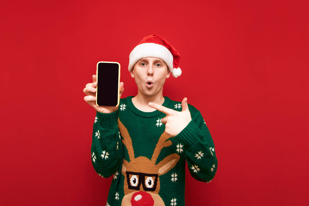 Surprised guy in santa hat and christmas sweater shows his hand on smartphone screen and looks into camera isolated on red background. Shocked guy shows black screen of smartphone into camera. - Foto, imagen