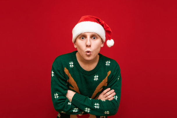 Portrait of surprised young man in santa hat and warm christmas sweater looking into camera with shocked face on red background. Photo of surprised guy in christmas clothes - Photo, Image