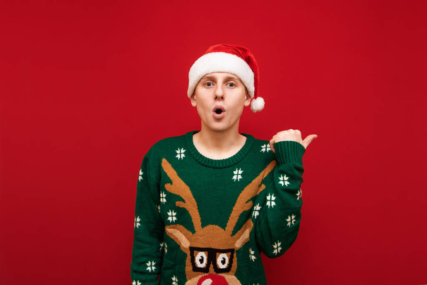Surprised young man in christmas hat and sweater isolated on red background, looks into camera shocked face and shows thumbs up at copyspace. Shocked Christmas guy points his hand to a blank space - Foto, Imagem