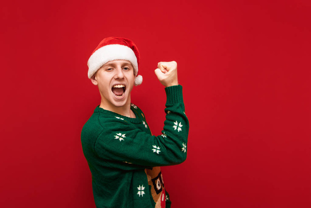 Portrait of joyful young man in christmas clothes, wearing santa hat and sweater, rejoices in victory on red background. The guy won at Christmas, he is happy. Isolated, New Year. - Photo, image