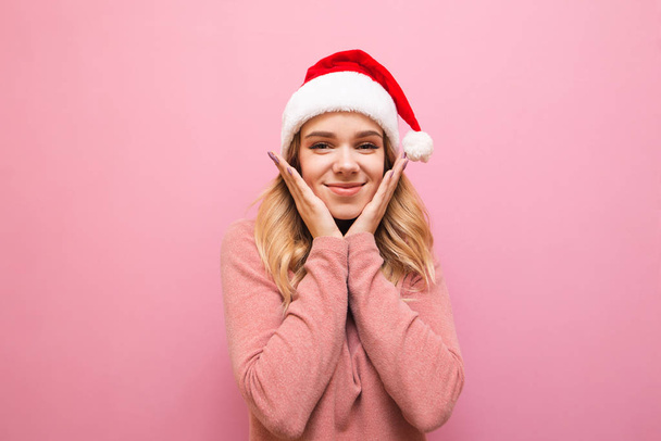 Портрет смешной девушки в рождественской шляпе на розовом фоне, улыбающейся мило и смотрящей в камеру. Изолирована. Симпатичная Санта девушка в розовом свитере позирует. Концепция x-mas
. - Фото, изображение